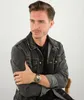 A man wearing a denim jacket and a black t-shirt, sitting and looking to the side. He is wearing a gold watch on his left wrist. Podgląd zdjęcia produktu