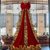 A large red bow is draped over a decorated Christmas tree, with gifts piled at the base. The scene is set indoors, possibly in a hotel lobby or a large room with a window view. Podgląd zdjęcia produktu