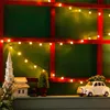 A Christmas-themed display on a shelf, featuring a miniature church, a small evergreen tree, and a white car carrying a Christmas tree, all illuminated by string lights. Voir les images du produit