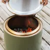 A close-up shot of a green fire pit with glowing embers and flames inside, with a silver lid being held above it. Produktbild-Vorschau