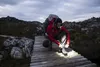 A person is tying their shoe on a wooden bridge in a rocky, mountainous area. Podgląd zdjęcia produktu