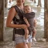 A woman is holding a baby in a carrier near a body of water, possibly under a pier. Anteprima dell’immagine prodotto