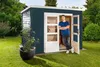 A man leaning against the open door of a dark blue wooden shed in a garden. Produktbild-Vorschau