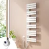 A white electric towel radiator is mounted on a light pink wall next to a window with greenery visible outside. A small digital controller is attached to the radiator. Voir les images du produit