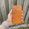 A hand holding a brown leather phone case in front of a wooden fence. Produktbild-Vorschau