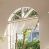 A sheer curtain with tassels hangs in an arched window, with trees and a building visible outside. Produktbild-Vorschau