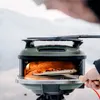 A person is using a portable pizza oven outdoors in a snowy environment. Produktbild-Vorschau