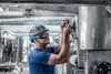 A worker in a blue shirt and safety gear is working on industrial machinery. Produktbild-Vorschau