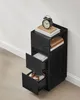 A black nightstand with drawers sits against a white wall and light wood floor. Náhled obrázku produktu
