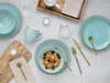 A flat lay of a turquoise dinnerware set with gold trim, including plates, bowls, and a mug, arranged with cutlery, a wooden cutting board, and a bowl of cereal with berries. Produktbild-Vorschau