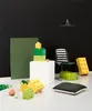 A collection of LEGO-style storage boxes and decorative items arranged on a white floor against a black wall. Produktbild-Vorschau