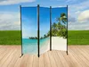 A three-panel room divider with a tropical beach scene printed on it, set up on a wooden deck with a grassy field and sky in the background. Produktbild-Vorschau