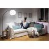 A woman sits on a couch in a living room, holding a smartphone. The room is decorated with framed pictures on the wall and a floor lamp. Produktbild-Vorschau
