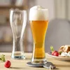 Two beer glasses, one filled with beer and foam, and the other empty, sit on a wooden table with radishes and a pretzel with dip in the background. Produktbild-Vorschau
