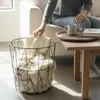A person's hand reaches into a wire basket filled with blankets, next to a wooden side table with a mug and book. Produktbild-Vorschau