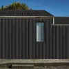 A close-up view of a dark gray corrugated metal building exterior with a small window and a section of roof visible against a clear blue sky. Náhled obrázku produktu