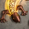 A close-up view of a decorative gecko-shaped wall light, made of metal with ornate cutouts and illuminated from within. Produktbild-Vorschau