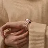 A close-up shot of a person's wrist wearing a rose gold metal watch band, with their other hand adjusting it. Produktbild-Vorschau