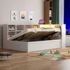A white bed with storage and a lifted mattress is shown in a bedroom setting with artwork on the wall and a bookshelf. Produktbild-Vorschau