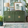 A green sideboard with decorative brass accents is placed in a room with light blue walls. It is decorated with two table lamps, books, and framed botanical prints. A potted plant is on the left, and a large potted plant is on the right. A patterned rug is Voir les images du produit