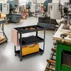 A three-tiered black metal tool cart with various tools and storage containers, placed in a workshop environment with machinery in the background. Náhled obrázku produktu