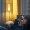 A woman sits in a chair, looking at her phone, with a floor lamp behind her. Anteprima dell’immagine prodotto