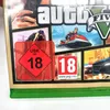 A close-up collage of images from the Grand Theft Auto V game cover, featuring various scenes and characters, with age rating labels prominently displayed. Produktbild-Vorschau