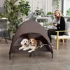 A woman sits on a chair on a patio next to a raised dog bed with a canopy, where two dogs are resting. There are plants and outdoor furniture in the background. Anteprima dell’immagine prodotto