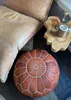 A close-up view of a leather pouf and a portion of a leather chair, with a wooden decorative item in the background. Podgląd zdjęcia produktu