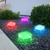 Four cube-shaped solar lights with different colored lights (red, purple, green, blue) are placed in a garden bed with dark gravel and surrounded by plants. Produktbild-Vorschau