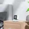 A small, white digital clock with a pixelated display sits on a wooden nightstand next to a bed. Náhled obrázku produktu