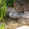 A small solar-powered fountain operating in a garden pond with surrounding rocks and plants. Produktbild-Vorschau