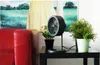 A black fan sits on a black table next to two potted plants, with a red couch and a painting in the background. Voir les images du produit
