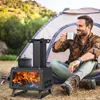 A man is sitting by a tent, drinking from a mug, with a portable wood stove in the foreground. Náhled obrázku produktu