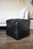 A black leather ottoman sits on a wooden floor in front of a white couch. Podgląd zdjęcia produktu