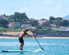 A man is paddleboarding on a body of water with a town and hills in the background. Produktbild-Vorschau