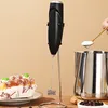 A black milk frother stands upright next to a stainless steel pitcher on a wooden surface. To the left, a dessert with white cream and green toppings sits on a plate. A spoon with white powder is held over the pitcher. Produktbild-Vorschau