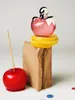 A still life arrangement featuring a perfume bottle, a candy apple, lemon slices, and a wooden block. Anteprima dell’immagine prodotto