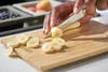 A person is slicing a banana on a wooden cutting board. Voir les images du produit