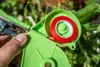 A hand is holding a green plant tying tool with red tape inside, with greenery in the background. Náhľad obrázku produktu
