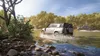 Two off-road vehicles driving through a shallow river surrounded by lush green trees and foliage. Produktbild-Vorschau