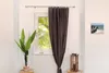 A curtain rod is shown mounted above a window, with a dark brown curtain drawn to the side. A potted plant is on the left, and a decorative lantern with a candle is on the right. A small framed picture of a beach scene hangs on the wall to the right. Produktbild-Vorschau