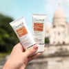 A hand holding two tubes of Guinot skincare products with a blurred background of a building and sky. Produktbild-Vorschau