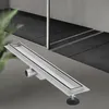 A silver linear shower drain with black rubber seals is shown in the foreground, angled diagonally. The background shows a modern bathroom with dark gray tiled flooring and a glass shower enclosure. Náhľad obrázku produktu