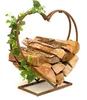 A heart-shaped metal log holder decorated with artificial ivy, filled with firewood, on a white background. Produktbild-Vorschau