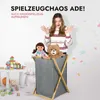 A young woman is standing behind a laundry basket filled with toys, with a surprised expression. There are toys on a table to the left and confetti in the air. Produktbild-Vorschau