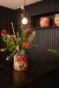 A colorful floral arrangement in a patterned vase sits on a dark countertop, with a light fixture above and decorative vases on a shelf in the background. Produktbild-Vorschau
