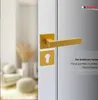 A close-up of a gold matte door handle and keyhole escutcheon set mounted on a white door, with a blurred interior scene in the background. Produktbild-Vorschau
