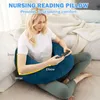 A woman is sitting on a bed with a U-shaped pillow around her, holding a phone. Produktbild-Vorschau