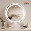A round LED mirror is displayed on a vanity table in a bedroom setting. Podgląd zdjęcia produktu